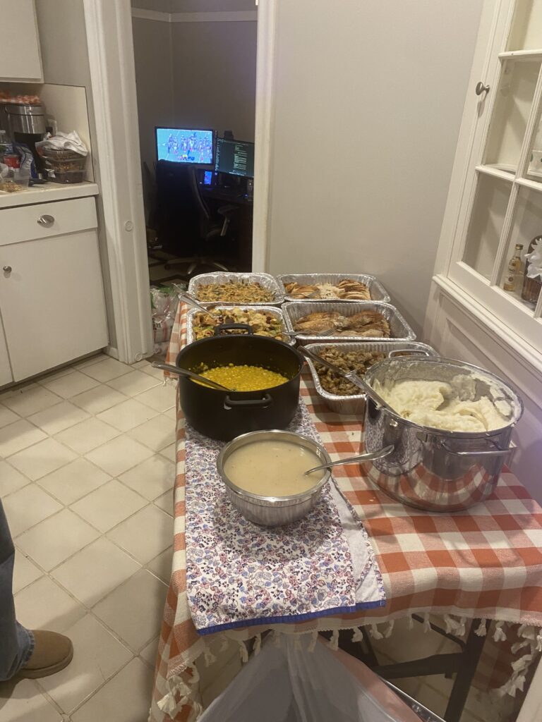 Tisch mit typischen Thanksgiving-Gerichten, im Hintergrund das Wohnzimmer mit laufendem Fernseher und einem Footballspiel